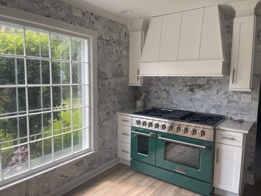 Modern kitchen featuring a teal gas range, white cabinets, and marble backsplash with large window.