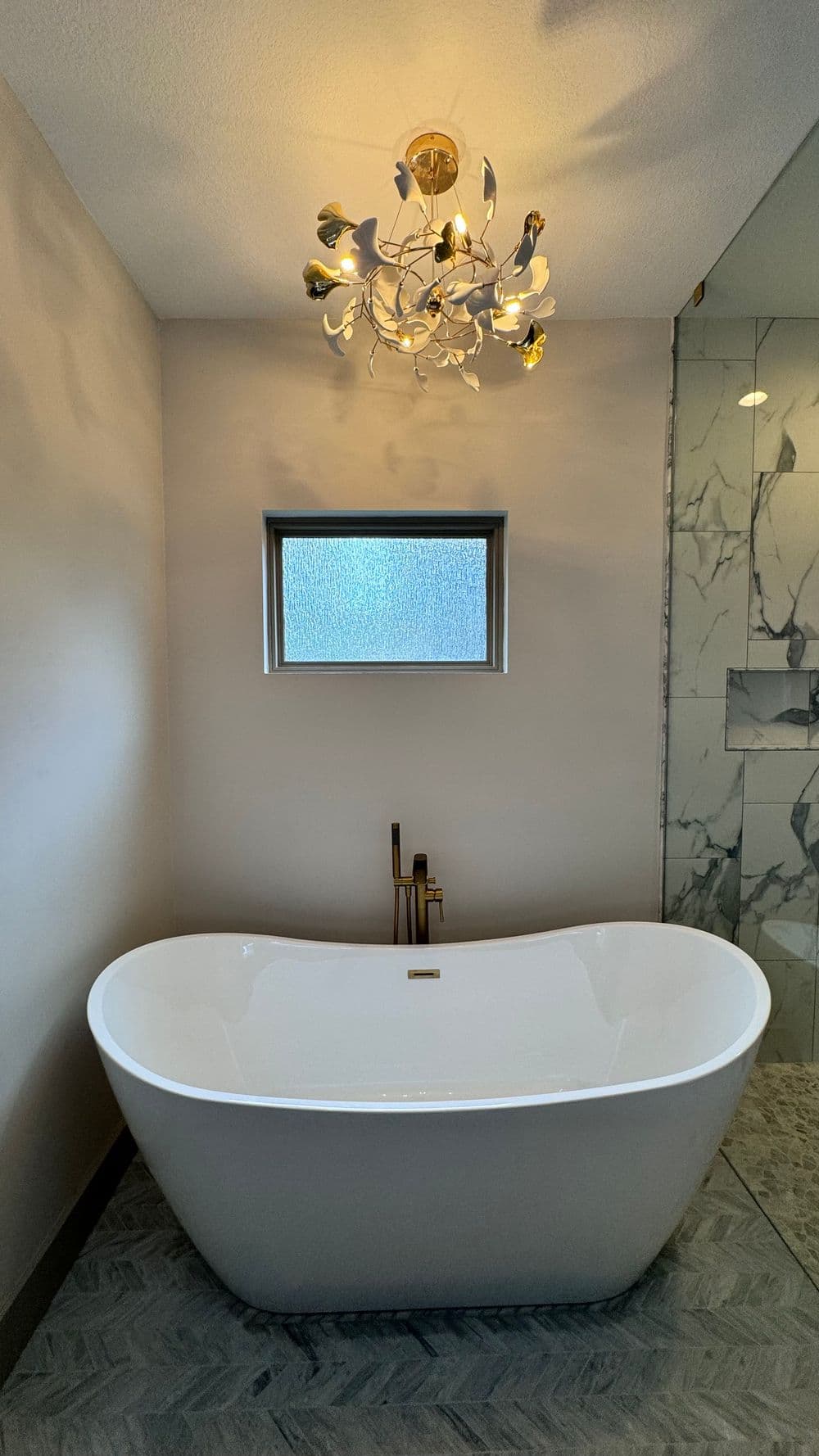 Modern bathroom with freestanding white tub, elegant chandelier, and marble accents.