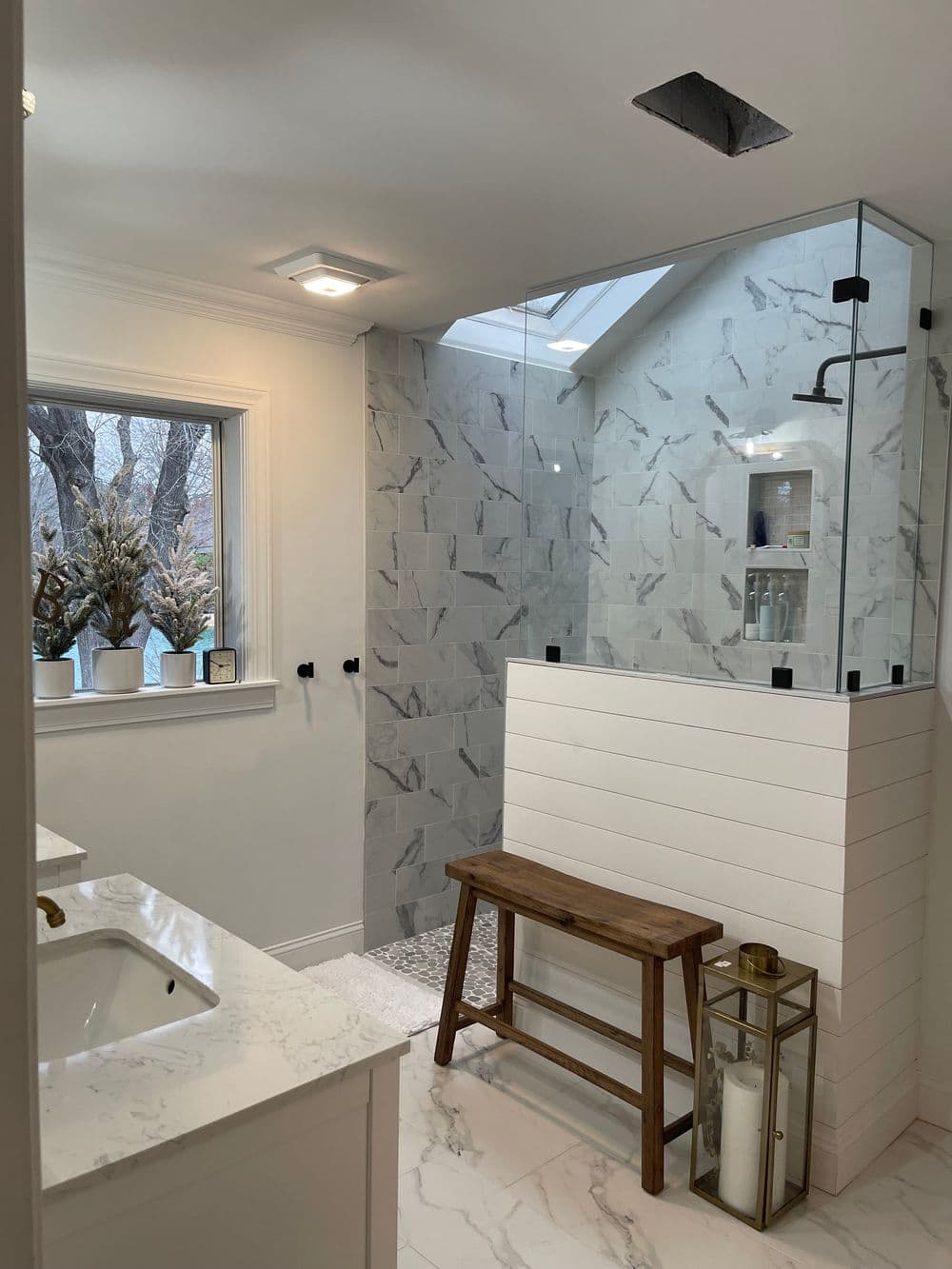 Modern bathroom featuring a glass shower, marble tiles, and a wooden bench under natural light.